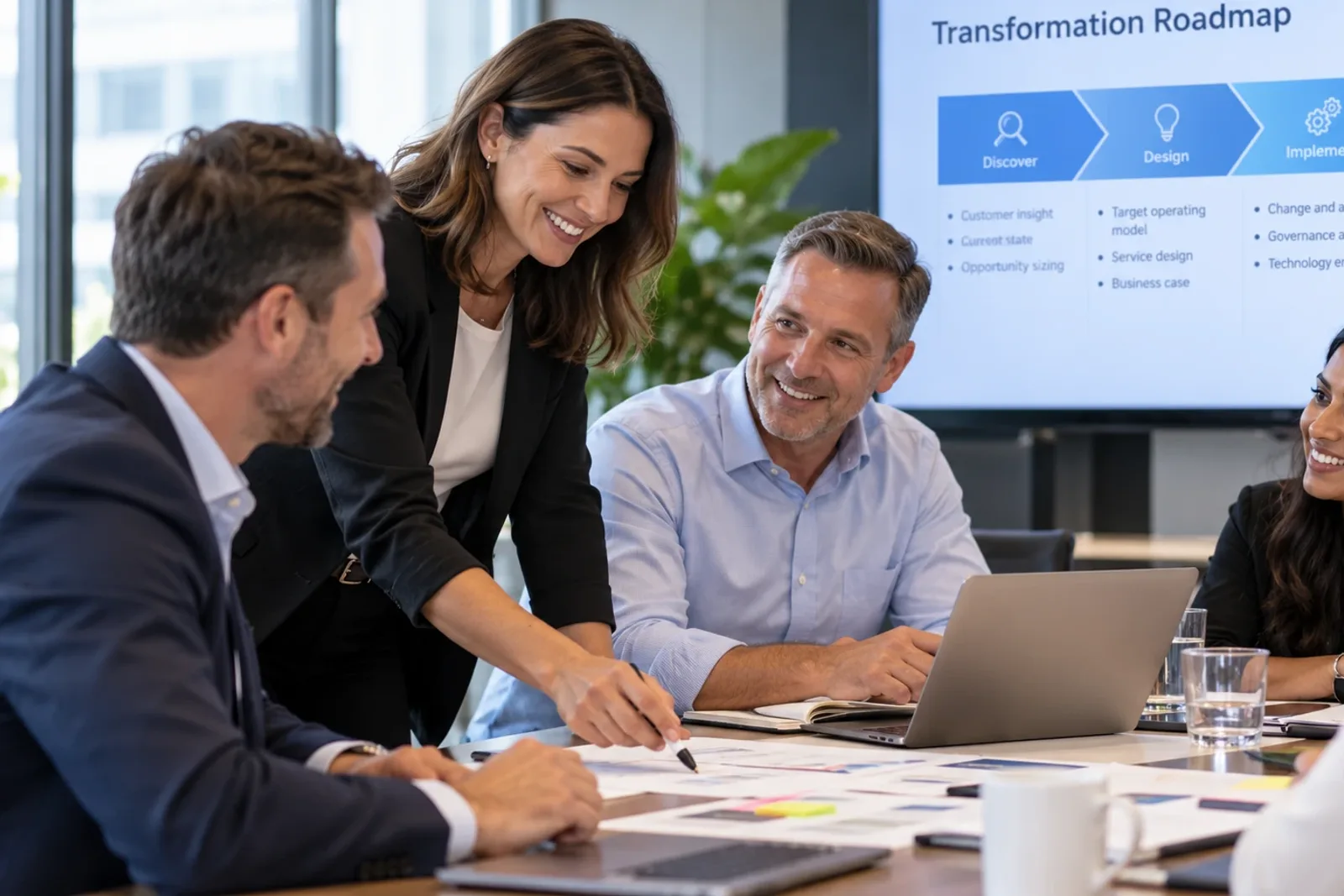 A consultant points to a plan on the table while colleagues smile, with a transformation roadmap on the screen behind them.