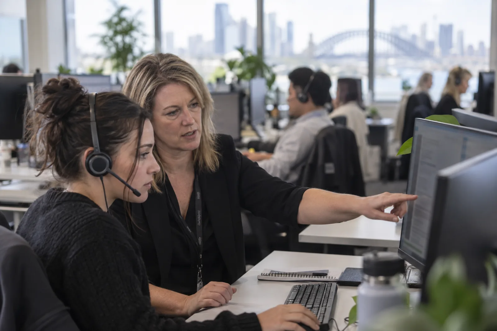 A contact centre team leader points to a passage on an agent’s screen while the agent, wearing a headset, follows along.